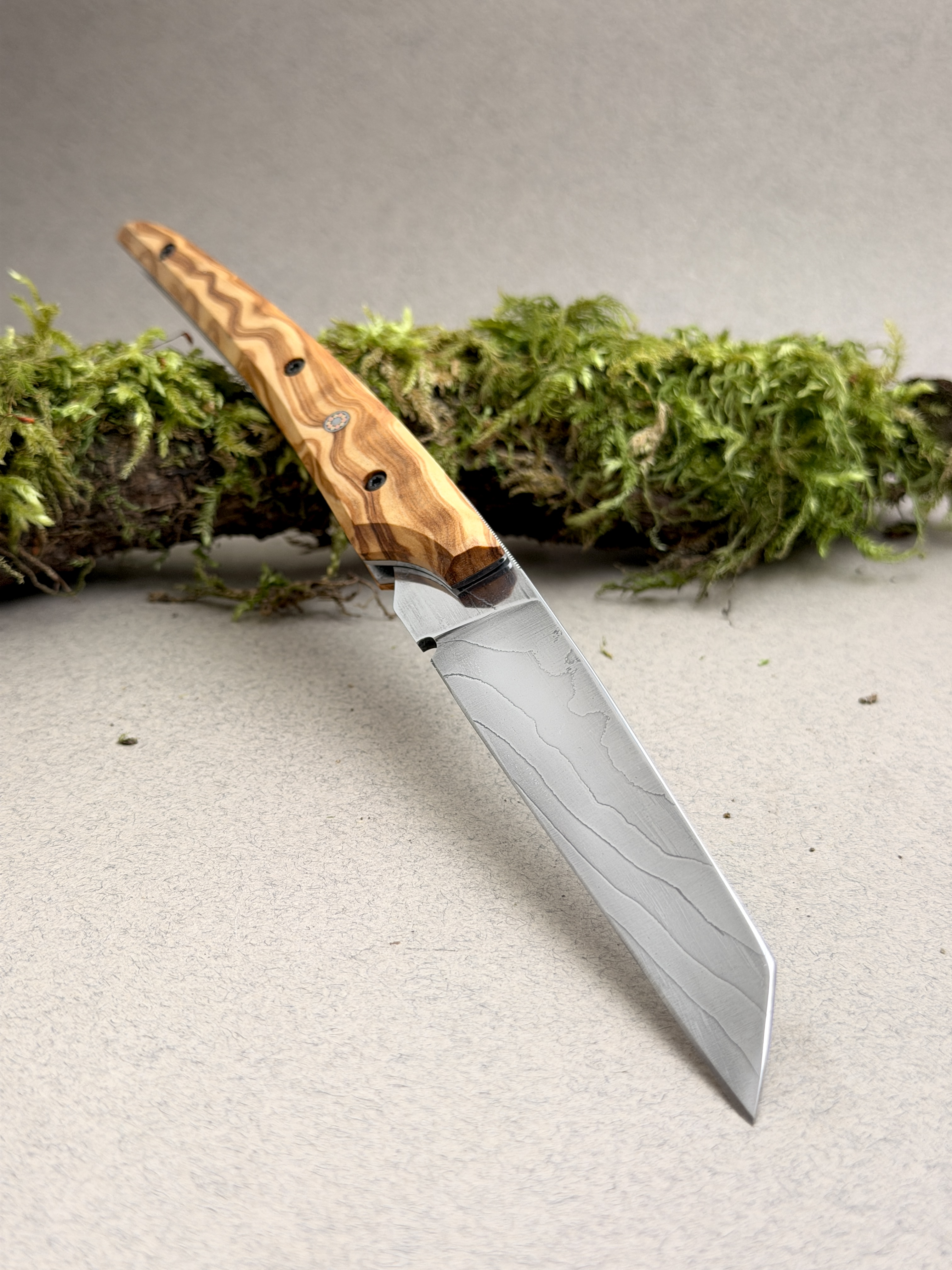 Handmade FPF Creations knife shown in full, with a wooden handle and a patterned blade, resting in front of a moss-covered branch.